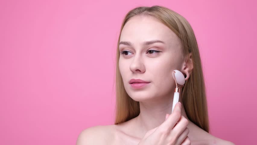 Beautiful young woman giving facial massage with stone roller on pink background. Concept of skin care and aging prevention. | Shutterstock HD Video #1111934099