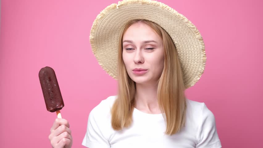 Happy young woman in straw hat eating popsicle standing on pink background. Concept of summer, vacation and cold desserts in hot weather. | Shutterstock HD Video #1111934109