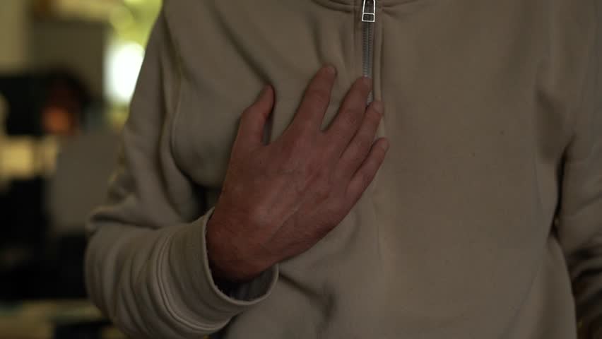 A man in a beige jacket in the room holds his hand on his heart. Flashing light in rhythm with heartbeat. Signs of stroke and heart disease. | Shutterstock HD Video #1111934995