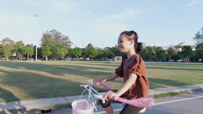 Backdrop of clear skies and lush greenery, this sporty Asian kid girl are riding bicycle for active outdoor living. Her winning smile and energetic spirit infuse the scene with joy and happiness. | Shutterstock HD Video #1111935261