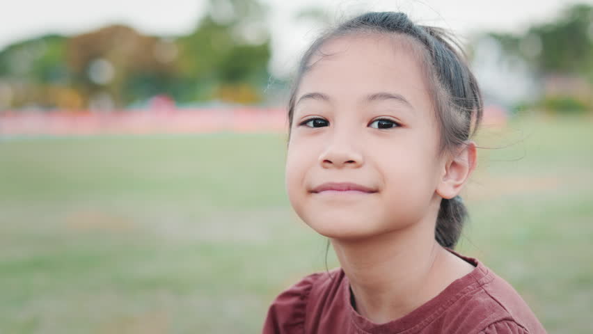 Lush greenery of a playground, close up with copy space of adorable Asian kid girl with innocence, positively, radiant smile and expressive face of happiness and beautiful summer vacation.