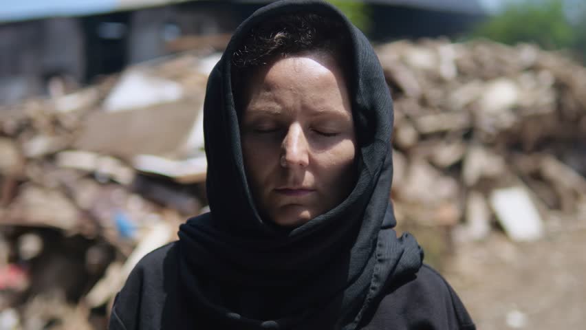 Grieving face of widowed woman with head covered with scarf. Grieving woman stands against backdrop of destroyed city, silently praying for dead relatives. Concept grief for those killed during war. Royalty-Free Stock Footage #1111935275