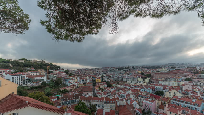Panorama showing aerial cityscape day to night transition from Miradouro da Graca viewpoint in Lisbon city after sunset. Dramatic clouds over historic houses with red roofs and evening illumination