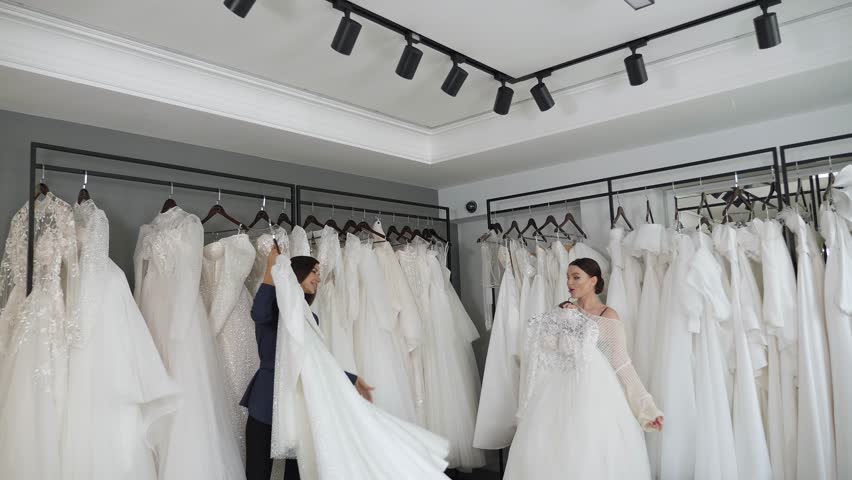 Two happy young women in a wedding salon, they hold in their hands wedding dresses that hang on hangers and try them on. Girlfriends from the wedding salon. Preparation for the wedding. Royalty-Free Stock Footage #1111938177