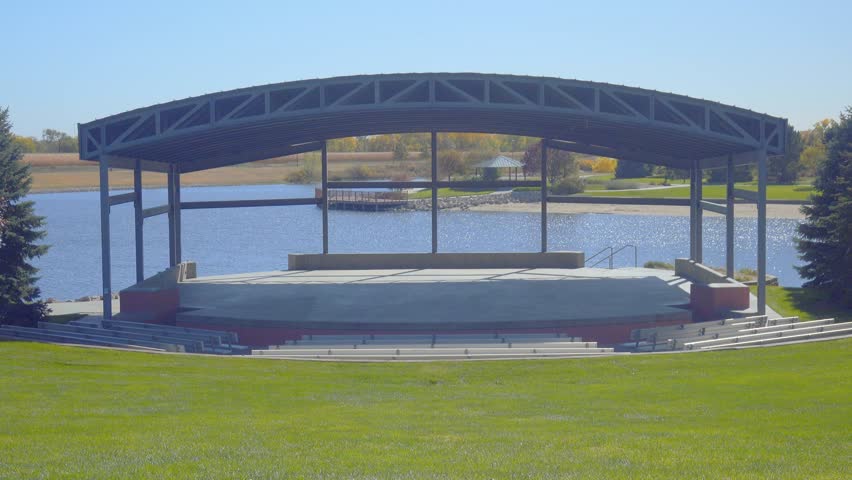 A man walks onto an empty stage at an outdoor park amphitheater on a beautiful sunny day.