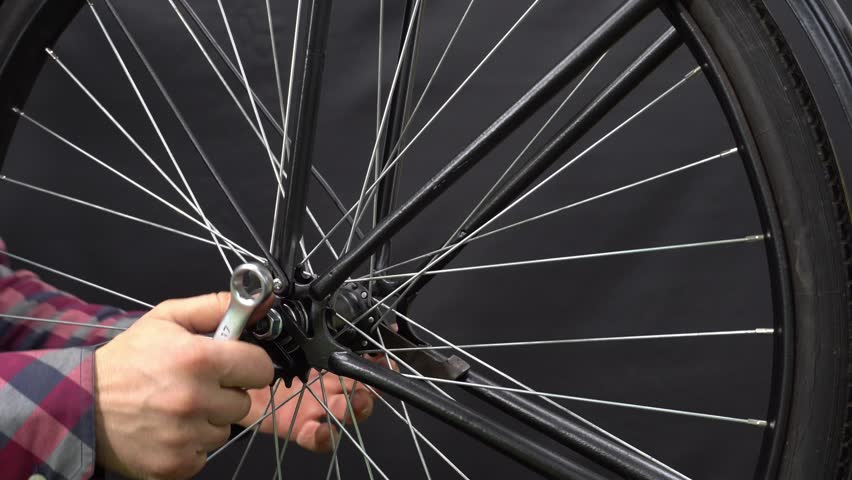 The mechanic installs the wheel on the bike. The wrench is in the hands of a mechanic. Black vintage bike in the workshop on a black background, studio light. bike details close-up | Shutterstock HD Video #1111942261