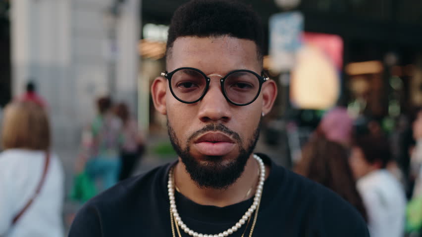 Charismatic young Black man exudes individuality and strong presence,he stands with arms crossed on a busy city street. His sharp gaze, framed by fashionable glasses illustrating self-assured attitude | Shutterstock HD Video #1111945105