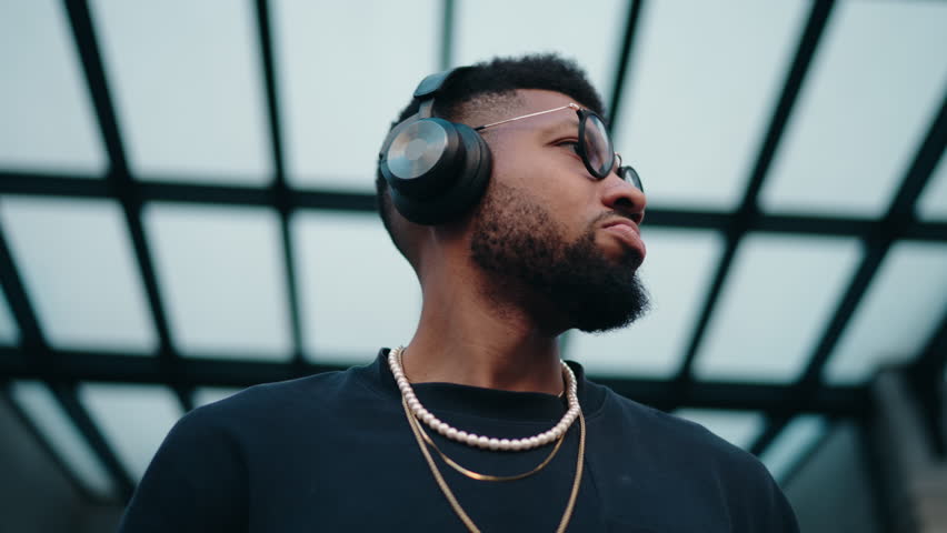 Young stylish dark skinned man listens intently to online playlist through his headphones, using modern smartphone apps, against a geometric backdrop. Musician in headphones feels the rhythm of sound | Shutterstock HD Video #1111945107