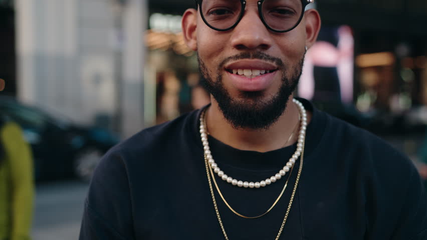 Portrait of young dark skinned man with radiant smile and stylish glasses.Trendy male wearing black t-shirt and sophisticated gold necklaces, dynamic city backdrop highlights his infectious positivity | Shutterstock HD Video #1111945113