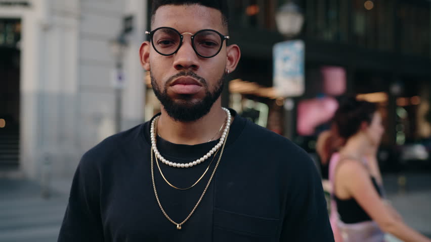 Close-up of young Black man with beard, wearing a black t-shirt and stylish glasses, adorned with chunky gold necklaces, set against a soft-focused urban background suggesting a lively city atmosphere | Shutterstock HD Video #1111945115