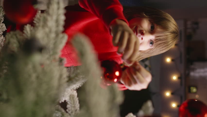 Cute kid boy, adorned in red pajamas, decks Christmas tree with cheerful red balls. Magic and joy of New Year celebration, capturing spirit of Merry Xmas. Cinematic AD. Vertical video. Amazing feast | Shutterstock HD Video #1111948921