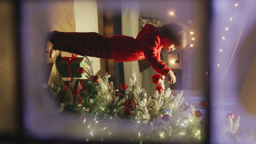 Cute boy in red pajamas decorating Christmas tree with red balls, outside view through snow frosted window panes. Vertical video, cinematic advertising. Waiting for a New Year's miracle, celebration | Shutterstock HD Video #1111948925