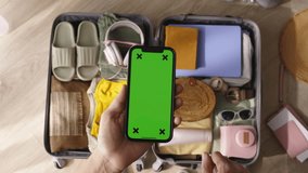 Top view smartphone with green chroma key in hand of woman. Checking list in special mobile app to see if everything is packed in suitcase advertising. Organized travel plans. Relaxation and adventure
