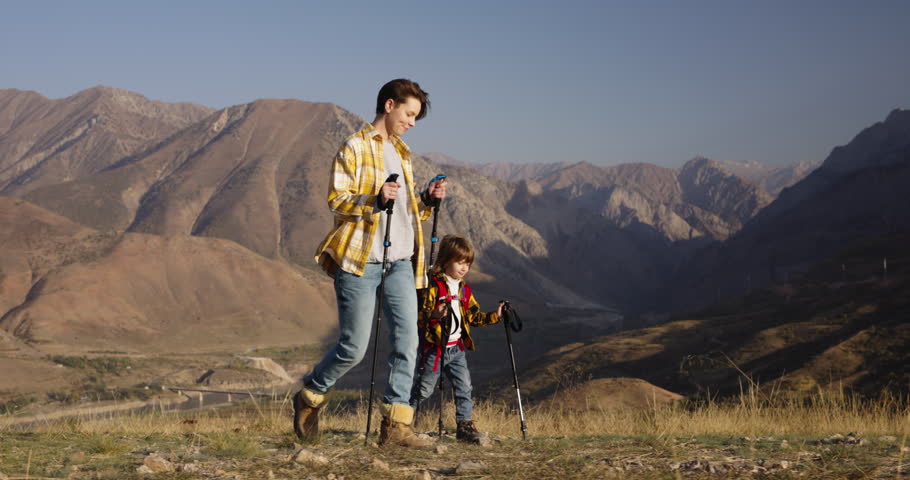 Mother and son conquer mountain, trekking poles backpacks. Step by step together mother and her son traverse rocky path. Reaching new heights, experience invigorating embrace of autumn sunlight | Shutterstock HD Video #1111948943