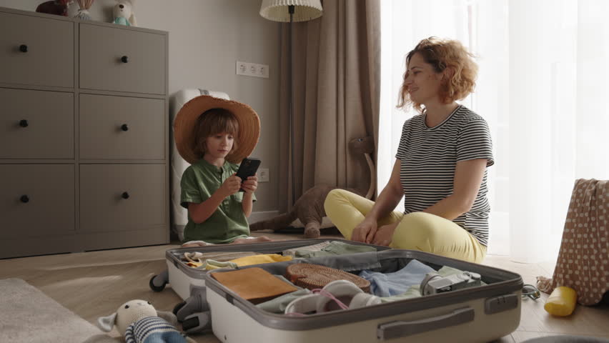 Mother and son engage in pre-trip preparations, side by side. Their precise packing and reliance on mobile checklist application demonstrates their strong collaboration for smooth journey | Shutterstock HD Video #1111948945