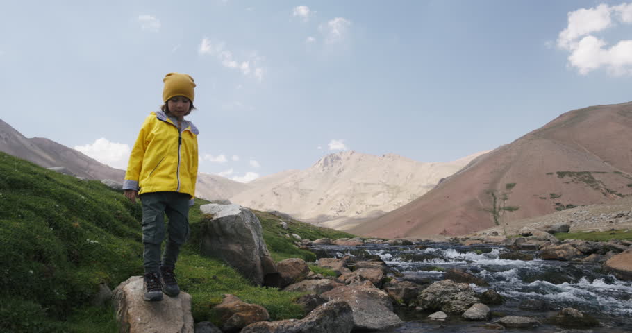 Child hiker crosses stream. Traveler boy walks through hilly terrain by river in search of adventure, exciting experiences. Active lifestyle, outdoor recreation in hilly area by river, walk by stream