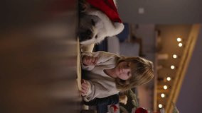 Charming little boy and white dog in red cap, crafting their Christmas wish list by twinkling tree. Cinematic AD. Boy whispers in dog ear. Santa anticipation. Vertical cinematic video, advertisement.