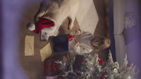 Little kid boy and his beloved dog decorating Christmas tree with red balls, outside view through snow frosted window panes. Creation of festive mood. Cinematic AD vertical video. Waiting for miracle