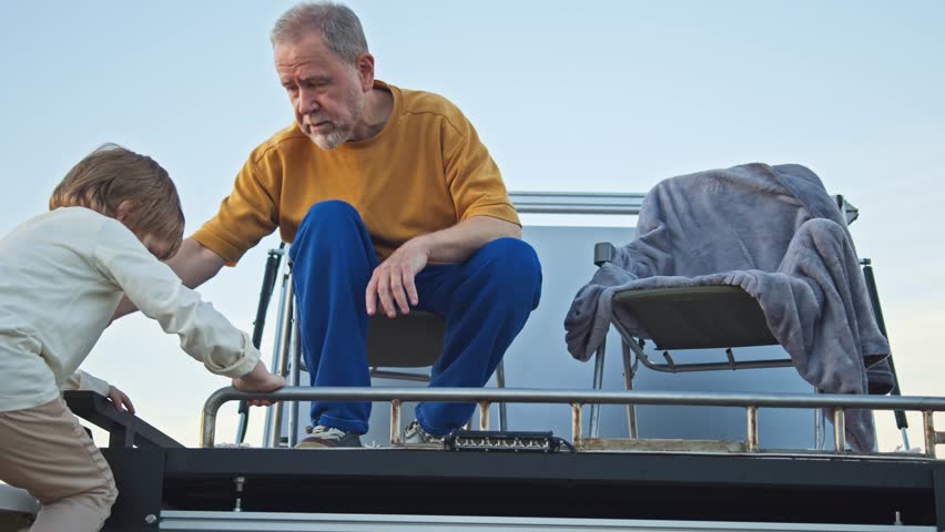 Grandpa assists boy onto camper rooftop for relaxation and sightseeing. Solar power station, energy panels | Shutterstock HD Video #1111949699