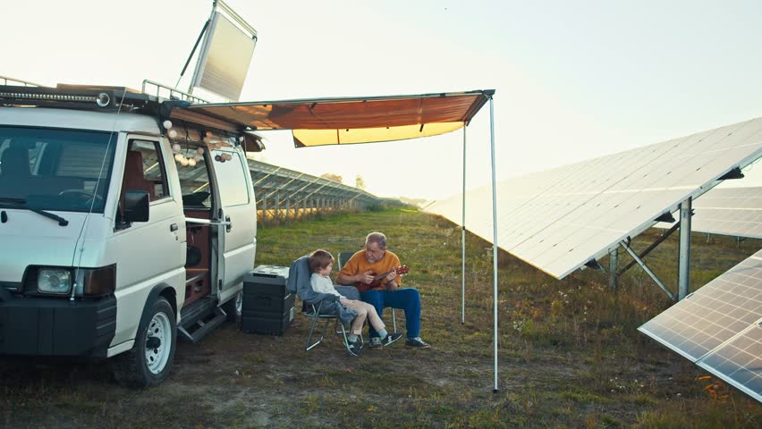 Grandfather and grandson sit outside their camper van near a solar panel field. Elder man plays the ukulele, teaching and bonding with the young boy during their outdoor vacation | Shutterstock HD Video #1111949715