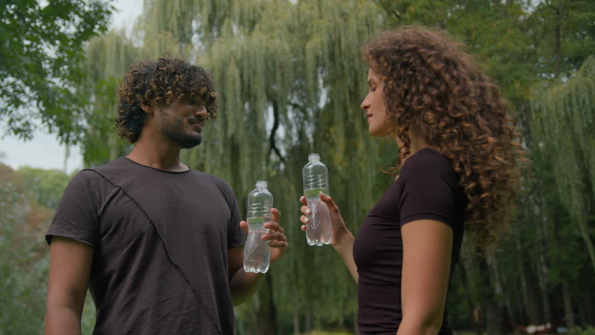 Diverse Indian man and Caucasian woman athletes drinking fresh water after city workout park outdoors young thirsty sport couple relaxing fitness break hydration vitamin drink recovery health wellness | Shutterstock HD Video #1111954705