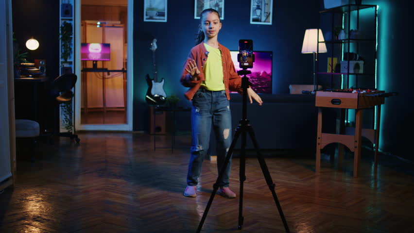 Cute youngster using mobile phone on tripod stabilizer to film dance tutorial for other children watching her online social media content. Girl recording video with smartphone targeting gen Z viewers | Shutterstock HD Video #1111955473