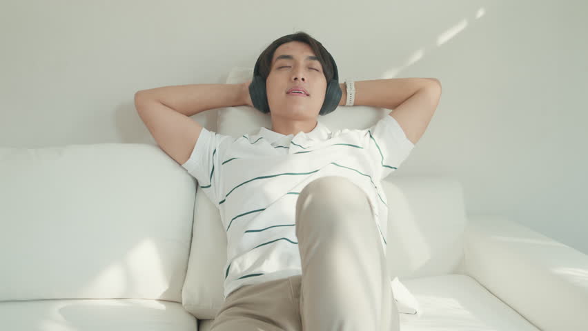 Youth attractive Asia male with casual cloth calm feeling lay down on sofa close eyes wear headphone listen music playlist relaxing at living room in cozy house. Lifestyle leisure at home concept. | Shutterstock HD Video #1111957823