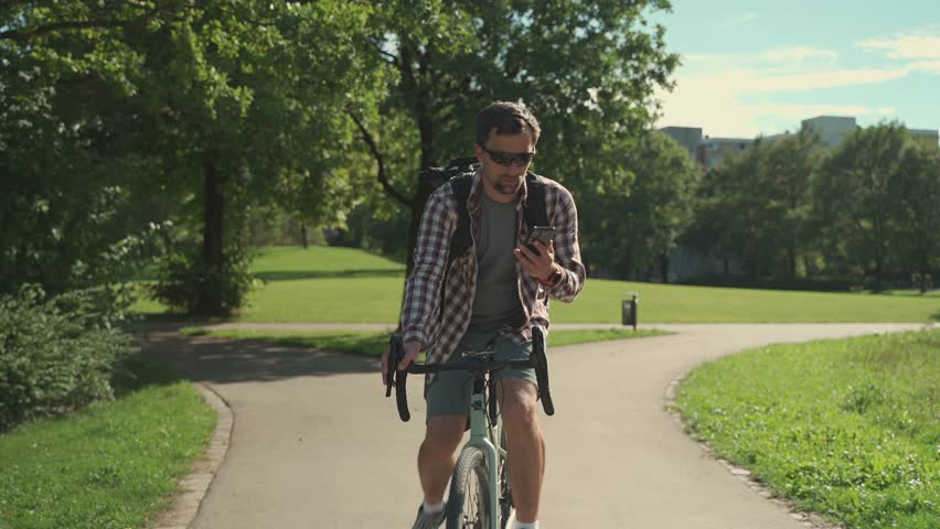Male cyclist with backpack rides bicycle path in park in summer and talks on his mobile phone. Active lifestyle and commuting on bicycle. Cyclist talking on telephone while cycling in the city park | Shutterstock HD Video #1111958905