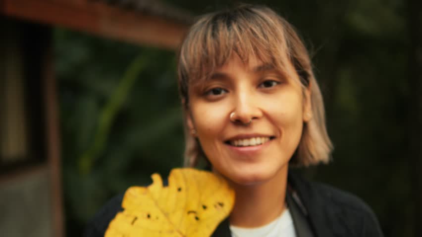 Portrait of a Young Woman Smiling at the Camera While Playfully Interacting with a Large Yellow Tropical Leaf in Front of Her Face, 4K Autumn Forest Lifestyle Video. | Shutterstock HD Video #1111960421