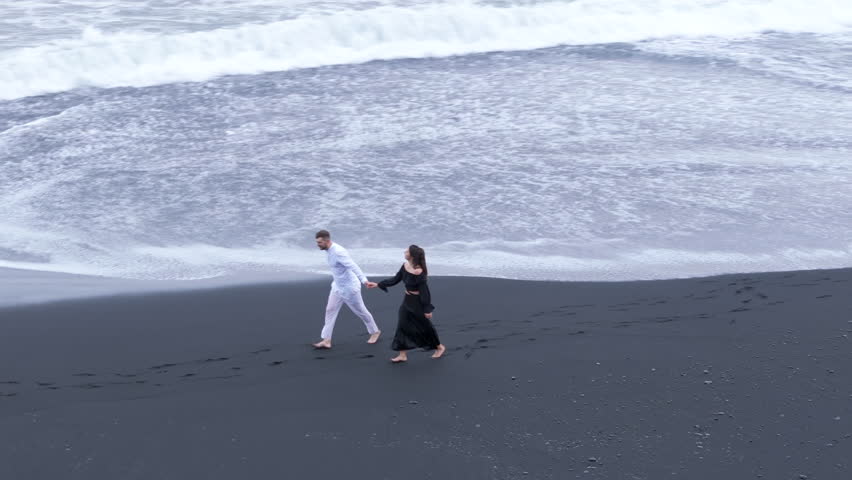 Loving young couple in black-and white clothes look at each other, rushing barefoot on black-sanded beach while foaming waves reach their feet. Iceland. High quality 4k footage Royalty-Free Stock Footage #1111961165