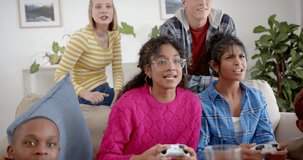 Happy diverse group of teenage friends sitting on couch and playing video games at home, slow motion. Gen z, friendship, hangout, technology and lifestyle, unaltered.