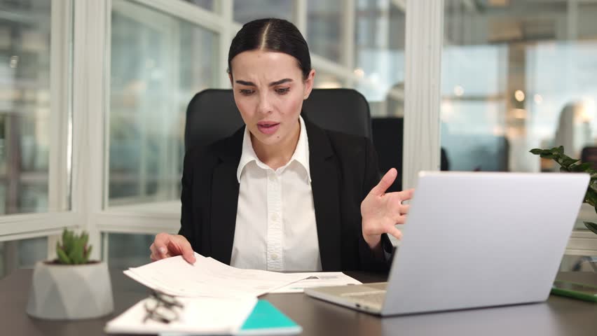 Worried office employee sitting at workplace and using modern laptop while reviewing failed reports indoor. Caucasian woman with braided hair holding paperwork and looking at bad statistics in office. | Shutterstock HD Video #1111967737