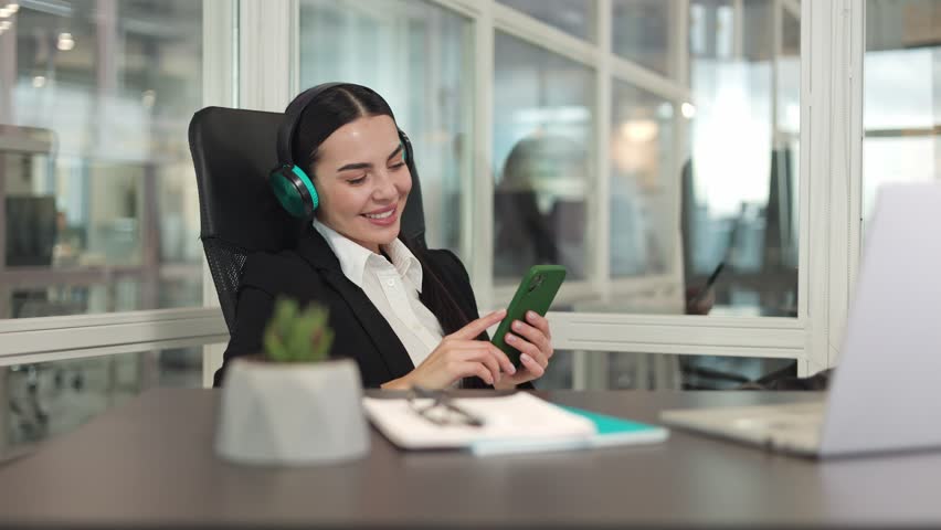 Relaxed business lady wearing black jacket sitting at table near modern laptop and holding smartphone while listening to music in headphones in office. Concept of break at work and favorite hobby. | Shutterstock HD Video #1111967749
