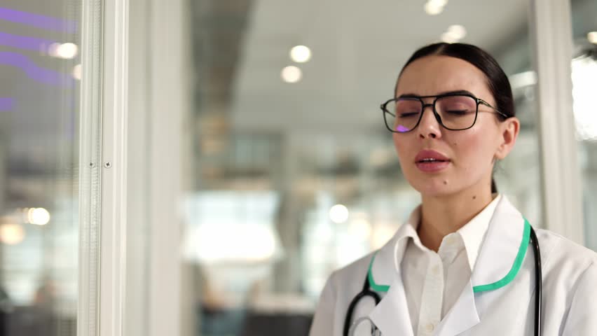 Sad female doctor closed in white medical uniform coming and leaning against wall while feeling headache. Caucasian professional therapist standing and putting hand to temple in modern hospital. | Shutterstock HD Video #1111967759