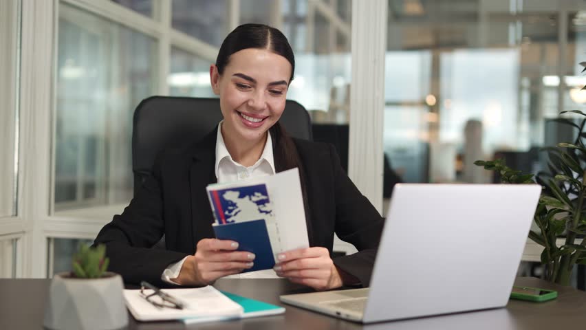 Optimistic woman employee having break at workplace and supporting head with hand while holding passport and tickets. Young brunette sitting with gadgets and thinking about vacation abroad in office. | Shutterstock HD Video #1111967761