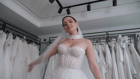 A luxurious female model in a wedding dress with beautiful makeup poses with a long veil in a modern wedding salon, she spins and looks at the camera. Stabilized image from below.