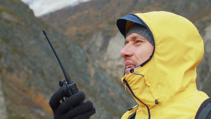 Man in jacket speaks on walkie-talkie standing in mountains. Side view. Search and rescue team worker reports information on portable radioset. Emergency response member use radio communication device Royalty-Free Stock Footage #1111976107