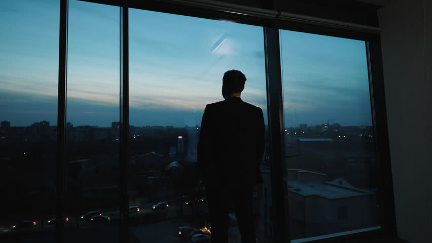 View from behind man in suit standing by panoramic windows in skyscraper, gazing at evening city. Businessman contemplates future while standing in office, observing sunset after completing his work. Royalty-Free Stock Footage #1111977437