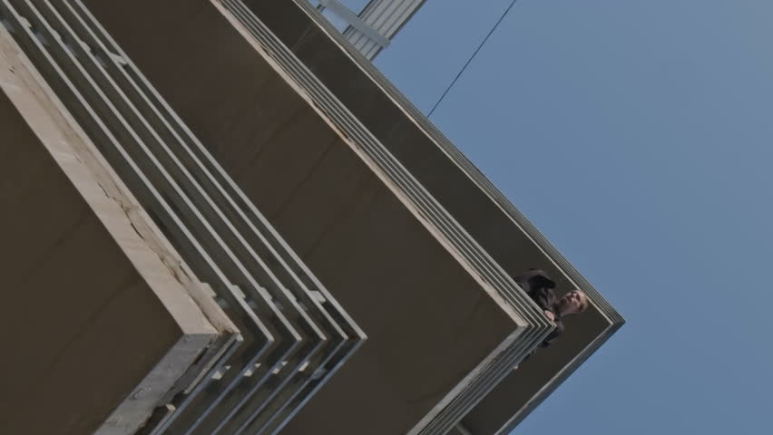 Vertical low angle shot of guy looking down on grown from height, sitting on railing of abandoned industrial site outdoors on clear sunny day | Shutterstock HD Video #1111977613