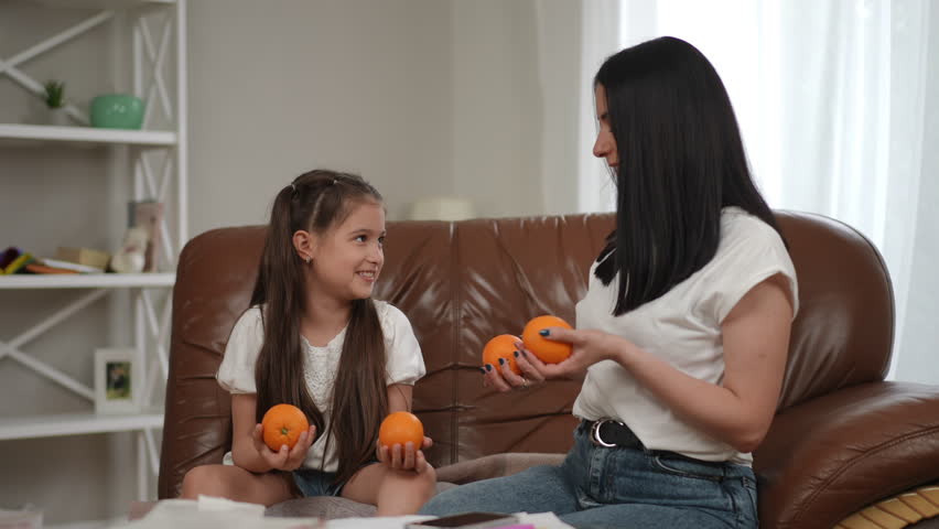 Fun time together. Mother and daughter sitting on the sofa in a modern house, juggling oranges