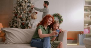 mom and girl make video call to far distance relatives on holidays to wish merry christmas. dad and son decorate christmas tree on background and wave hello