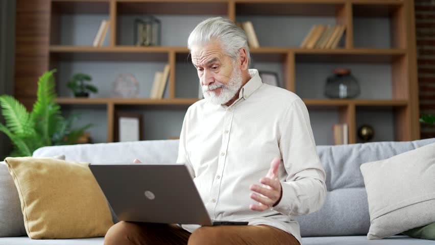 Confused senior old man with gray hair having difficulty using laptop sitting on sofa in living room at home. Worried puzzled mature male has problem complexity with work on application on computer | Shutterstock HD Video #1111982487