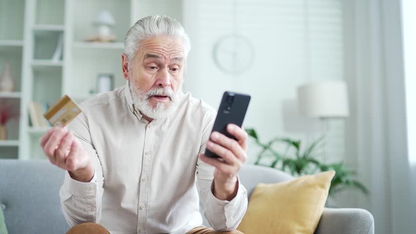 Frustrated elderly senior man discovered fraud while entering credit card number on smartphone sitting at home. Upset mature male became a victim of deception, money had been stolen from his account | Shutterstock HD Video #1111982511