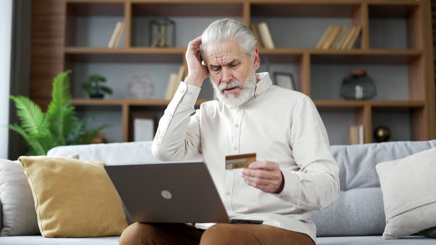 Frustrated elderly senior man discovered fraud while entering credit card number on laptop sitting on sofa at home. Upset mature male became victim of deception, money had been stolen from his account | Shutterstock HD Video #1111982515