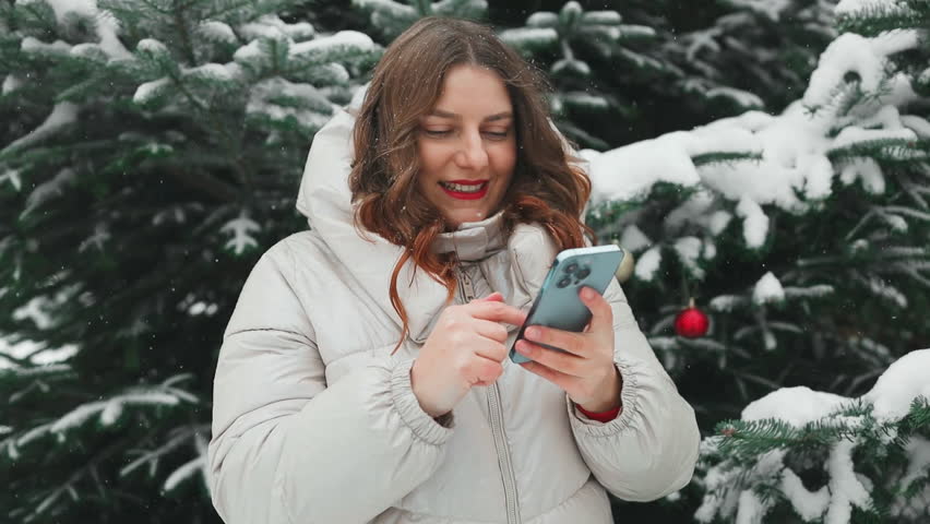 Young beautiful woman using her mobile phone at a snowy winter park. Closeup of female checking news and texting on her cellphone outdoor during cold winter season.  | Shutterstock HD Video #1111985847