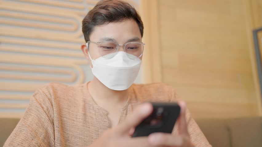 A happy man wearing a face mask sits typing a message on his smartphone. | Shutterstock HD Video #1111986475