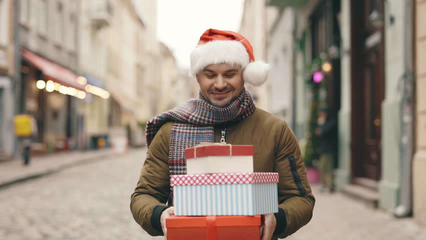 Cheerful Handsome Man Walking on Background of Decorated Building and Holding New Year's Gifts. Positive Bearded Guy with Holiday Presents Going Home | Shutterstock HD Video #1111992199