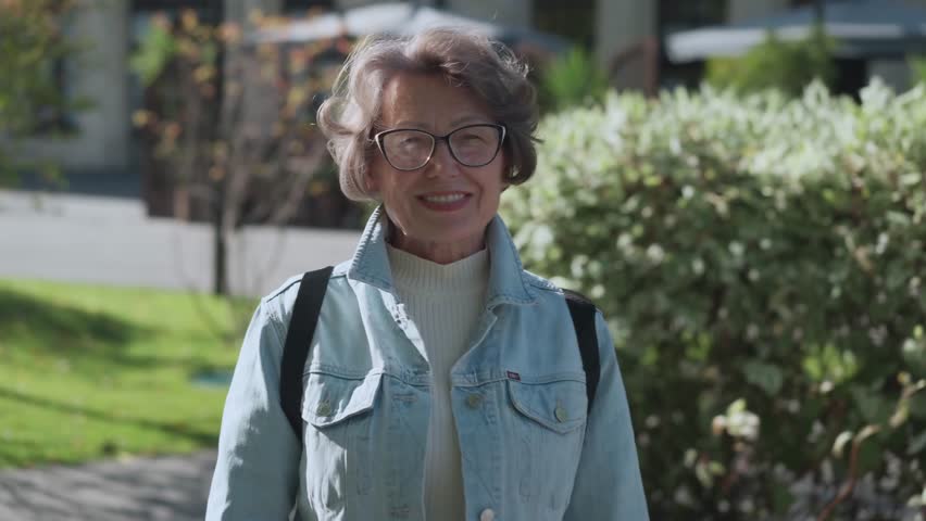 An elderly charming woman with shiny smile standing outdoors park. Portrait of pretty old lady in denim shirt at city center. Beautiful traveler female pensioner cheerful outside city | Shutterstock HD Video #1111993741