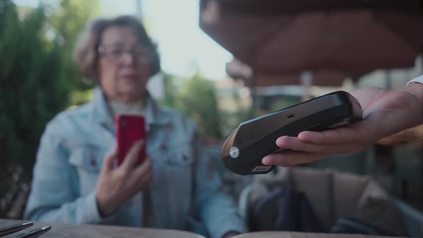 An elderly woman in denim shirt with cellphone contactless pay at restaurant. Old grandmother using smartphone nfc payment at cafeteria in city center. Progressive senior lady cashless pays for | Shutterstock HD Video #1111993761