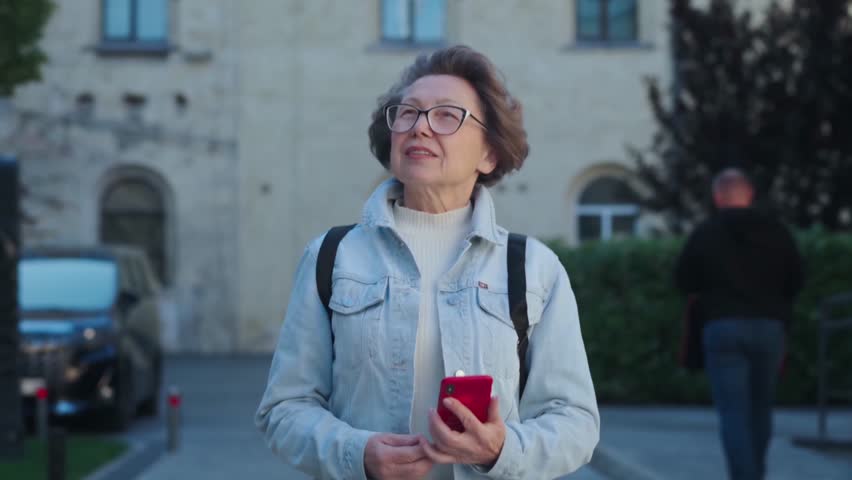 Old woman exploring old european city and using her phone. Pretty mature lady strolling with smartphone and looking around to find better location. Charming woman in windy weather traveling | Shutterstock HD Video #1111993785